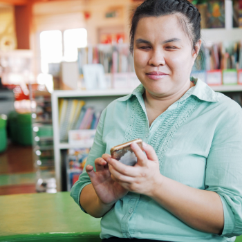 Smiling woman in a green shirt holding a smartphone, seated indoors with bookshelves and bright light in the background.